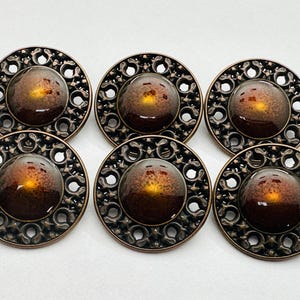 Peut inclure: Six boutons décoratifs ronds. Chaque bouton présente un motif filigrané de couleur bronze autour d'un dôme central en verre ambré. Ces boutons sont adaptés à la couture ou aux projets d'artisanat et ont un style vintage.
