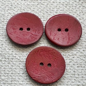 May include: Three round, dark red buttons with a textured, slightly uneven surface. Each button features two small holes in the center for attachment. The buttons have a matte finish and a consistent circular shape, arranged in a triangular formation.