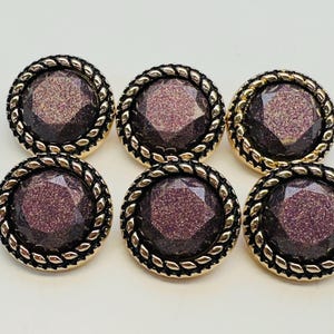 May include: Six round decorative buttons. Each button features a glittery purple centre, a gold-coloured rope-style border, and a black outer edge. The buttons are arranged on a white surface.