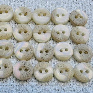 May include: Twenty round buttons with two holes, in a mix of off-white and tan. The buttons have a marbled effect. They are arranged in a grid pattern on a textured, light-coloured surface.