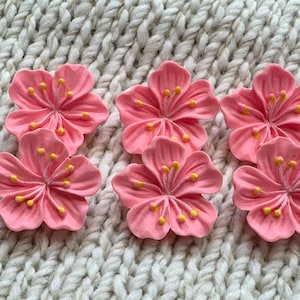 Puede incluir: Un conjunto de cinco adornos con forma de flor rosa, cada uno con cinco pétalos redondeados y un grupo central de pequeños estambres amarillos. Las flores están dispuestas en grupo, mostrando un diseño consistente con un acabado mate suave.