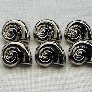 Puede incluir: Seis botones brillantes de color plateado con diseño de concha. Cada botón tiene un centro en espiral y una forma curva y redondeada. Los botones están dispuestos sobre una superficie clara y texturizada.