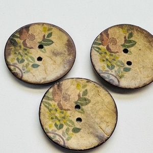Puede incluir: Tres botones redondos de madera con un diseño floral. Los botones tienen un fondo marrón claro con un patrón floral descolorido en rosa, amarillo y verde. Cada botón tiene dos agujeros para coser.
