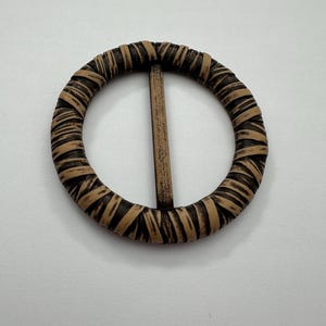 May include: A round belt buckle with a woven design in shades of brown and tan. The buckle has a central bar and a circular frame, suitable for belts or other accessories.