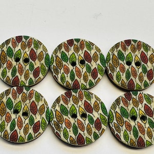 May include: Six circular wooden buttons featuring a leaf design in green, orange, and yellow hues. Each button has two black sewing holes. The buttons are displayed on a white background.