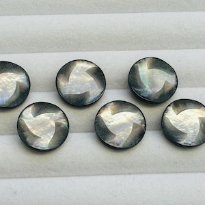 May include: Six circular, dark grey buttons with a mother-of-pearl design. Each button has a swirling, three-part pattern in the centre, creating a unique visual effect. The buttons are arranged in a neat row.