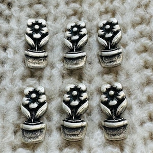 May include: Six small, silver-coloured flower pot charms. Each charm features a detailed flower with petals, leaves, and a pot. The charms are arranged on a textured, light-coloured surface.