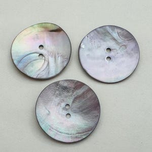 May include: Three iridescent, round buttons with two holes each. The buttons have a pearlescent sheen with swirls of gray, purple, and green. The buttons are made of mother of pearl and are suitable for sewing projects.
