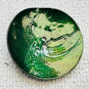 May include: A round, green decorative dish with a glossy finish. The dish has a textured surface and a small hole near the center. The color transitions from dark green to a lighter, almost iridescent green.