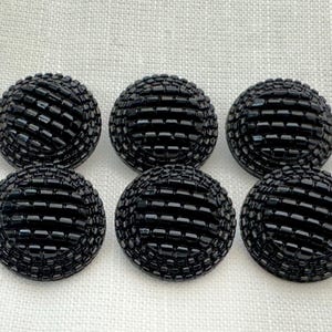 May include: Six round, black buttons with a textured, woven-like design. The buttons are evenly spaced on a light-coloured, textured fabric background. Each button has a slightly domed shape.