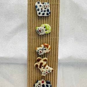 Puede incluir: Un conjunto de cinco botones de cerámica con forma de gato en varios colores y diseños, fijados a un soporte de cartón corrugado. Los gatos presentan detalles como manchas, rayas y rasgos faciales. Los botones son pequeños, decorativos y adecuados para proyectos de costura o manualidades.