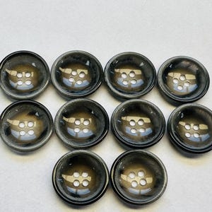 Puede incluir: Diez botones gris oscuro con un acabado brillante. Cada botón tiene una forma ligeramente abovedada y cuatro agujeros para coser. Los botones están dispuestos en un patrón ordenado sobre un fondo blanco.