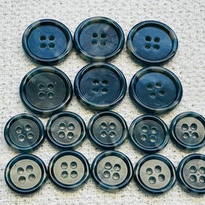 May include: A selection of dark teal buttons in various sizes. Each button features four holes and a marbled effect. The buttons are arranged on a white textured surface, showing their different diameters.