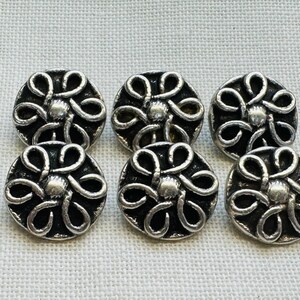 Peut inclure: Un ensemble de cinq boutons circulaires présentant un motif floral en métal argenté en relief avec six pétales en boucle entourant un point central, sur un fond noir profond en creux. Les boutons ont une finition métallique et ronde.