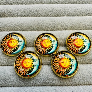 Puede incluir: Cinco cabujones redondos de vidrio con borde dorado con un diseño de sol y luna. El sol es amarillo con una cara sonriente y la luna es azul con forma de media luna. El fondo es negro con estrellas.