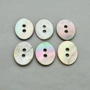 Può includere: Sei bottoni ovali in madreperla, ciascuno con due fori, disposti in due file da tre. I bottoni hanno una lucentezza iridescente, con colori rosa, verde e giallo. Lo sfondo è grigio.