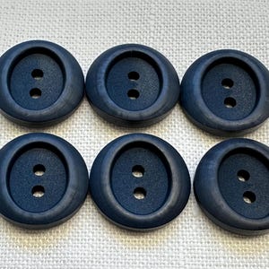 May include: Six navy blue, round buttons, each with two holes. The buttons have a slightly raised, rounded edge and a matte finish. They are arranged in two rows of three on a textured, off-white surface.