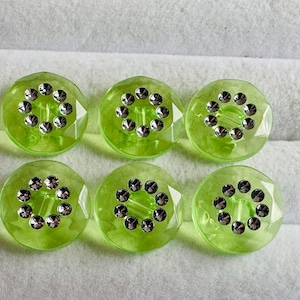 May include: Six round, lime green buttons with a faceted edge. Each button has a silver-colored, circular design in the center. The buttons are arranged on a white, textured surface.