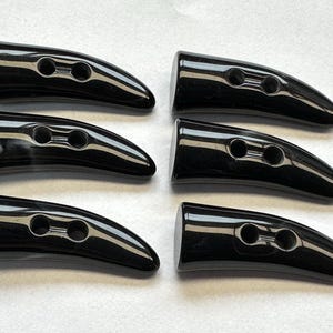 May include: Six glossy black horn-shaped buttons with two holes each. The buttons are arranged in two rows of three, with a smooth, curved design. These buttons are suitable for coats, jackets, or other garments.