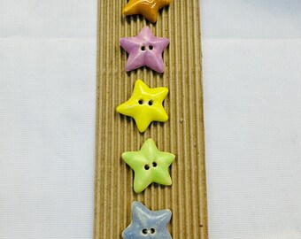 May include: Five star-shaped buttons in a row, each with two holes for sewing. The buttons are in shades of orange, purple, yellow, green, and blue. They are mounted on a brown, corrugated cardboard strip.