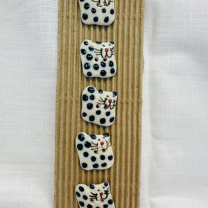 May include: Five ceramic cat-shaped buttons with black spots and red noses are arranged vertically on a corrugated cardboard strip. The buttons have a white base colour and are detailed with black spots and facial features.