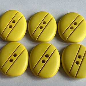 Może przedstawiać: Sześć jasnożółtych, owalnych guzików. Każdy guzik ma dwa otwory i dwa ukośne rowki. Guziki ułożone są w dwóch rzędach po trzy, eksponując ich design i kolor.