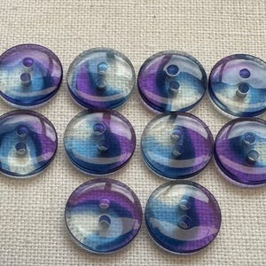 May include: Ten clear, round buttons, each with two holes. The buttons feature a swirl pattern in blue, purple, and white. They are displayed on a light beige fabric.