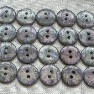 以下が含まれることがあります： 20個の虹色のグレーのボタンのコレクション。各ボタンには2つの穴があります。ボタンは、紫と緑の色合いを持つ真珠光沢のある光沢があります。明るいベージュ色の生地に格子状に配置されています。
