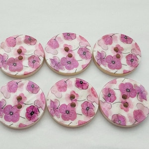 May include: Six round wooden buttons with a floral design. Each button features a white background with pink and purple flowers. The buttons have two holes for sewing and a light brown edge.