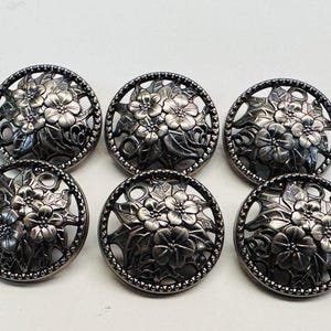 Puede incluir: Seis botones antiguos plateados con diseño floral. Cada botón presenta un arreglo floral detallado dentro de un marco calado circular. Los botones miden aproximadamente 2,5 cm de diámetro, ideales para proyectos de costura o coleccionismo.