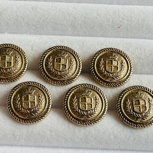 Może przedstawiać: Zestaw siedmiu antycznych, złotych guzików. Każdy guzik ma szczegółowy wzór herbu z koroną, tarczą i wieńcem laurowym. Guziki mają obramowanie w kształcie liny i są ułożone na białej powierzchni.