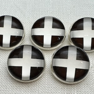 May include: Five round, silver-rimmed buttons with a dark brown background and a white cross design. The buttons are arranged on a light-colored, textured surface.