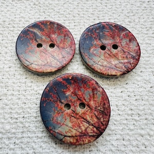 May include: Three circular buttons with a nature-inspired design. Each button features a print of red and orange tree branches against a blue and grey sky. The buttons have two holes for sewing and are arranged on a white, textured surface.