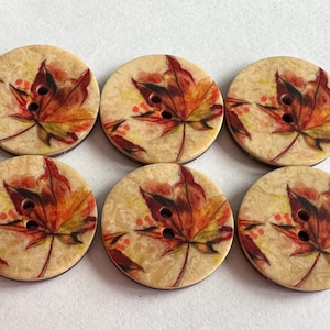 May include: Six round wooden buttons with a fall leaf design. The leaves are depicted in shades of red, orange, and yellow, with a light beige background. Each button has two holes for sewing.