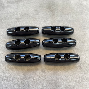 May include: Six black plastic toggle buttons with two holes each. The buttons are oval shaped and have a glossy finish.