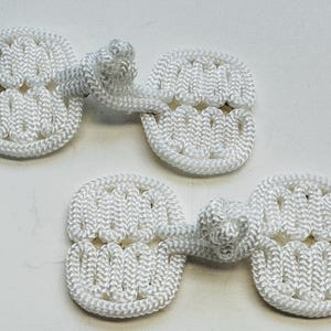 May include: Two white frog closures, a type of button, are shown against a white backdrop. Each closure comprises two rounded, connected sections with a knotted loop for fastening. The closures are made of woven cord.