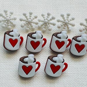 May include: Six white buttons shaped like mugs filled with brown liquid and white marshmallows, each featuring a red heart on the front. Above the mugs are five white snowflake-shaped buttons with rounded tips arranged in a horizontal row.