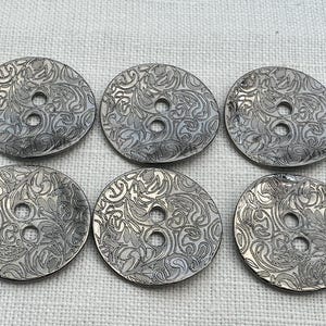 May include: Six silver buttons with a detailed floral design. Each button has two holes for sewing. The buttons are circular and have a slightly raised edge, creating a textured appearance.