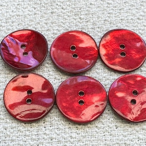 May include: Six red, round buttons with two holes each. The buttons have a glossy, iridescent finish, with shades of red and pink. The buttons are arranged on a textured, off-white surface.