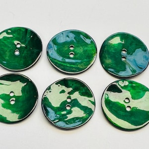 May include: Six round, glossy buttons with a marbled green and white design. Each button has two holes for sewing and a black edge. The buttons are arranged on a white surface.