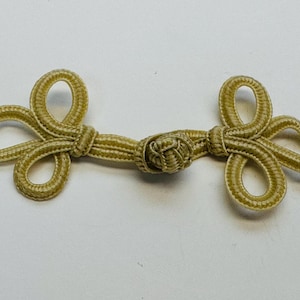 May include: A gold-colored frog closure, a decorative fastener commonly used on garments. The closure features a central knot with looped ends on either side, creating a symmetrical design. The closure is made of a woven material.