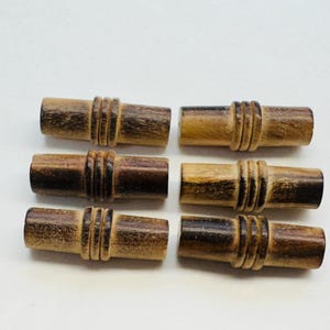 May include: Six wooden toggle buttons with a brown and dark brown striped pattern. The buttons are cylindrical and have three grooves around the center.