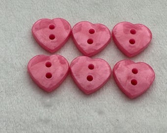 May include: Six pink heart-shaped buttons with two holes each. The buttons are made of a shiny, pearlescent material.