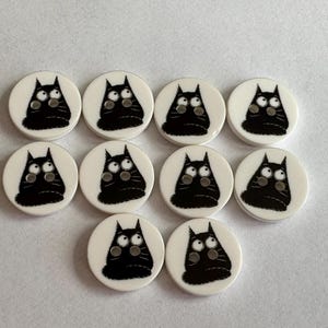 May include: Ten white, circular buttons featuring a black cat design. Each button shows a black cat with large eyes and a surprised expression. The cat has a fluffy tail and is centered on the button.