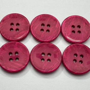May include: Six round, magenta buttons with four holes each. The buttons have a marbled effect and are arranged in two rows of three on a white surface. These buttons are suitable for sewing and craft projects.
