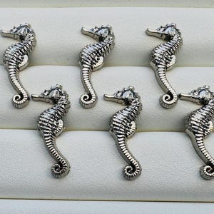 May include: Six silver seahorse charms displayed on a white surface. Each charm features detailed scales and a curled tail. The seahorses are positioned in various directions, creating a dynamic arrangement.
