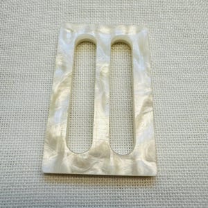 May include: A rectangular belt buckle featuring a pearlescent off-white finish with a marbled texture. The buckle has two vertical, elongated oval cutouts positioned side-by-side, creating a three-bar design for threading a belt strap through.