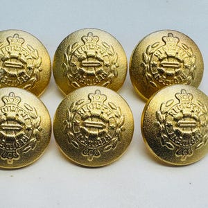 Puede incluir: Seis botones de metal de color dorado con un diseño en relieve que presenta una corona y una letra "R" estilizada.
