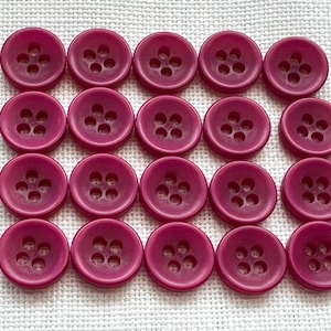 May include: A grid of twenty round, magenta-colored buttons arranged in four rows of five. Each button features a smooth, slightly concave surface with four centered holes for sewing, maintaining a uniform circular shape and consistent color.