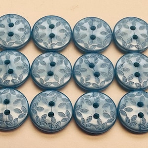 Puede incluir: Doce botones redondos de color azul claro con un diseño de hojas en los bordes. Cada botón tiene tres agujeros para coser. Los botones están dispuestos en tres filas de cuatro.
